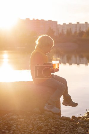 Téléchargez les photos : Belle enfant blonde d'âge préscolaire, garçon, assis dans le parc le matin lever du soleil, profiter du temps d'automne, boire du thé et profiter du soleil - en image libre de droit