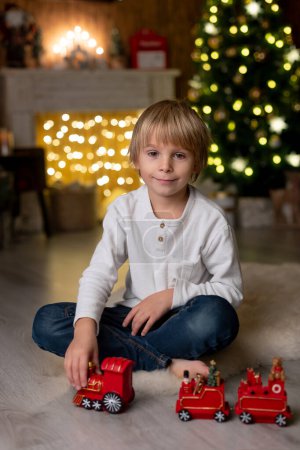 Photo for Beautiful blond child, young school boy, writing homework for school in a decorated home, drinking milk and eating cookies. Ligths and knitted toys around - Royalty Free Image