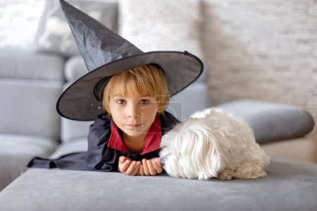 Photo for Cutel little child, boy in halloween costume, lying on the couch with pet dog, watching TV and cuddling - Royalty Free Image