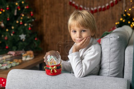 Photo for Beautiful blond child, young school boy, writing homework for school in a decorated home, drinking milk and eating cookies. Ligths and knitted toys around - Royalty Free Image