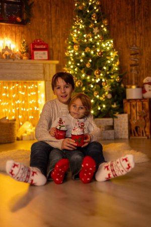 Foto de Dulces niños lindos increíbles, hermanos varones, abrazándose en casa frente a la chimenea en Navidad, amor en familia - Imagen libre de derechos