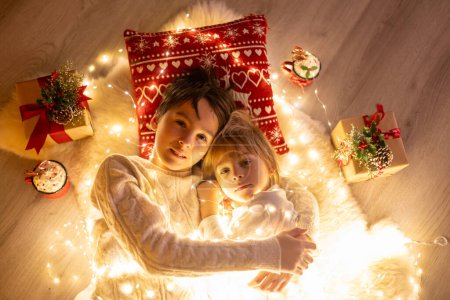 Téléchargez les photos : Doux enfants mignons étonnants, garçons frères, câlins à la maison devant la cheminée à Noël, amour en famille - en image libre de droit