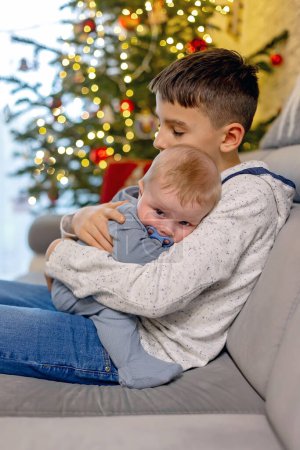 Photo for Preteen child, boy brother, hugging his little newborn sibling at home, sitting on the couch at home on christmas - Royalty Free Image