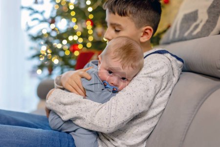 Téléchargez les photos : Enfant préadolescent, garçon frère, embrasser son petit frère nouveau-né à la maison, assis sur le canapé à la maison le jour de Noël - en image libre de droit