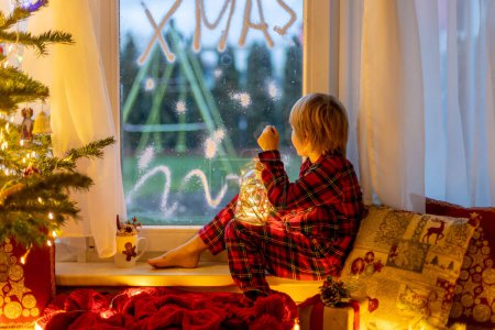 Photo for Cute child, sitting on a window, looking outdoors for Santa Claus, eating cookies - Royalty Free Image