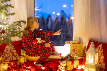 Photo for Cute child, sitting on a window, looking outdoors for Santa Claus, eating cookies - Royalty Free Image