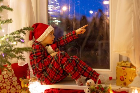Téléchargez les photos : Mignon enfant, assis sur une fenêtre, regardant à l'extérieur pour le Père Noël, manger des cookies - en image libre de droit