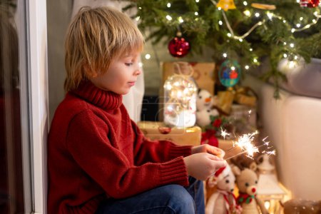Foto de Lindo niño, sosteniendo chispeante en la víspera de año nuevo en casa, sentado frente al árbol de Navidad, sonriendo - Imagen libre de derechos