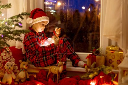Téléchargez les photos : Mignon enfant, assis sur une fenêtre, regardant à l'extérieur pour le Père Noël, manger des cookies - en image libre de droit