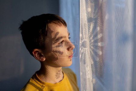 Téléchargez les photos : Portrait artistique d'un adolescent, debout à côté d'une fenêtre, ombres de rideaux tombant sur son visage, faire des ombres intéressantes, portrait d'art - en image libre de droit