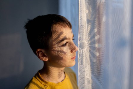 Téléchargez les photos : Portrait artistique d'un adolescent, debout à côté d'une fenêtre, ombres de rideaux tombant sur son visage, faire des ombres intéressantes, portrait d'art - en image libre de droit
