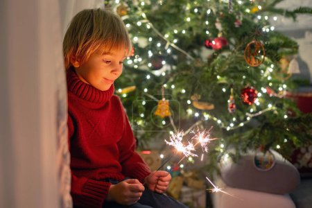 Foto de Lindo niño, sosteniendo chispeante en la víspera de año nuevo en casa, sentado frente al árbol de Navidad, sonriendo - Imagen libre de derechos