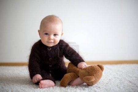 Foto de Pequeño bebé lindo, vestido con osito de peluche marrón hecho a mano en general, jugando en casa en el dormitorio soleado - Imagen libre de derechos