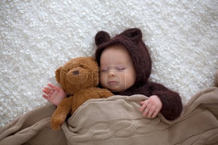 Téléchargez les photos : Petit garçon doux, vêtu d'ours en peluche doux brun tricoté à la main dans l'ensemble, dormant confortablement à la maison dans une chambre ensoleillée avec beaucoup d'ours en peluche autour de lui - en image libre de droit