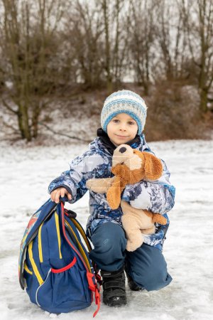 Téléchargez les photos : Enfant mignon, écolier avec sac à dos scolaire et chien en peluche après l'école dans la neige, l'hiver - en image libre de droit
