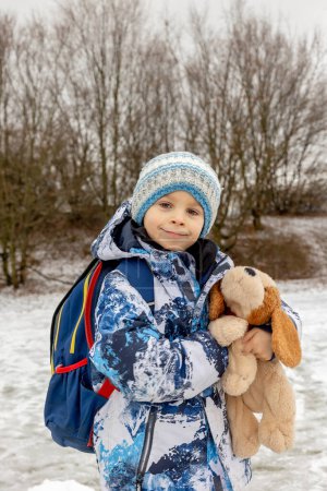Foto de Lindo niño, niño de escuela con mochila escolar y perro de peluche después de la escuela en la nieve, invierno - Imagen libre de derechos