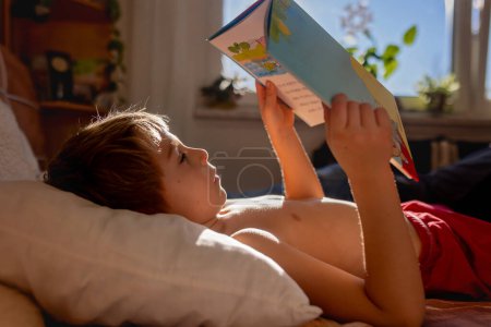 Téléchargez les photos : Enfant mignon, garçon, couché dans le lit avec livre, lecture le matin, temps ensoleillé en hiver - en image libre de droit