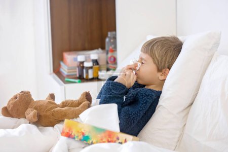 Téléchargez les photos : Enfant malade, tout-petit garçon couché au lit avec une fièvre, se reposant à la maison avec un ours en peluche, éternuant dans un mouchoir - en image libre de droit