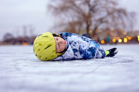 Foto de Niño con casco, niño, patinando durante el día en el lago congelado, acostado sobre su espalda después de caer - Imagen libre de derechos