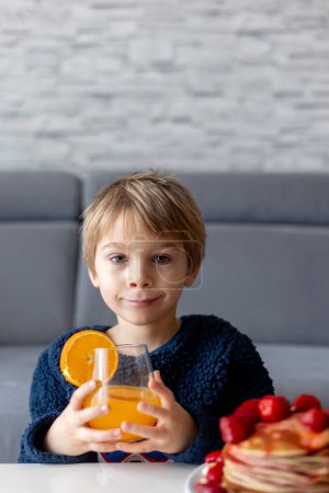Foto de Niño dulce, niño, comiendo panqueques americanos con fresas y plátanos, cubierto con jarabe y bebiendo jugo de naranja fresco - Imagen libre de derechos