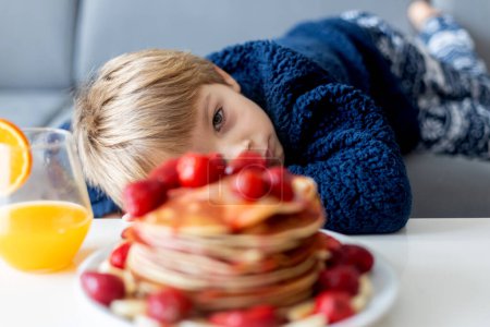 Photo for Sweet toddler child, boy, eating american pancakes with strawberries and bananas, topped with syrup and drinking fresh orange juice - Royalty Free Image