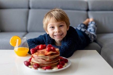 Téléchargez les photos : Enfant tout-petit sucré, garçon, mangeant des crêpes américaines aux fraises et aux bananes, garni de sirop et buvant du jus d'orange frais - en image libre de droit