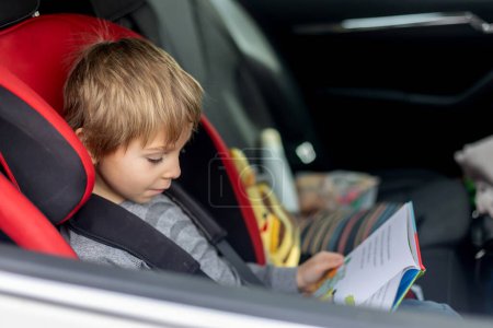 Foto de Lindo niño en silla de coche para niños, viajando en coche y leyendo un libro - Imagen libre de derechos
