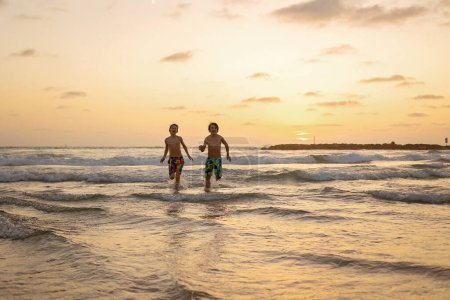 Téléchargez les photos : Heureux adolescents garçons, courir et jouer sur la plage au coucher du soleil, éclabousser l'eau et sauter sur le sable. Tel Aviv, Israël - en image libre de droit