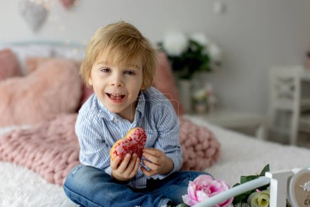 Téléchargez les photos : Mignon petit enfant blond, garçon d'âge préscolaire, manger beignet rose en forme de coeur, fait pour la Saint-Valentin, fond Valentin - en image libre de droit