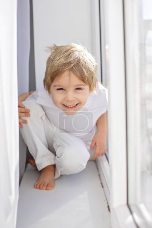 Téléchargez les photos : Petit garçon heureux mignon, enfant en chiffons blancs, souriant à la caméra joyeusement, jouant à côté d'une fenêtre avec des rideaux blancs - en image libre de droit