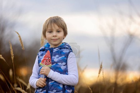 Foto de Lindo niño, hermoso chico rubio, comiendo piruleta de sandía en el parque al atardecer, hermoso clima de primavera al aire libre - Imagen libre de derechos