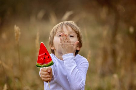 Photo for Cute child, beautiful blond boy, eating watermelon lollipop in the park on sunset, beautiful spring weather outdoors - Royalty Free Image