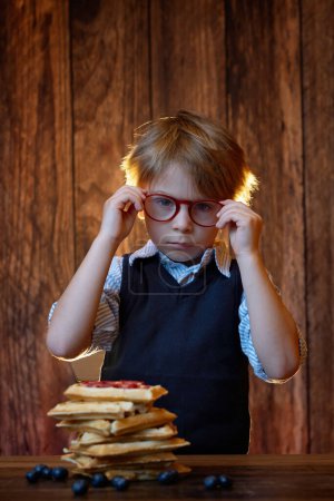 Téléchargez les photos : Enfant tout-petit sucré, garçon, mangeant des gaufres belges aux framboises, aux bleuets, aux noix de coco et au chocolat pour le petit déjeuner, plan studio - en image libre de droit