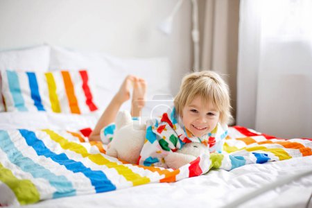 Téléchargez les photos : Bébé enfant heureux, garçon blond avec peignoir coloré, assis dans le lit avec jouet en peluche après la baignoire, souriant, literie colorée - en image libre de droit