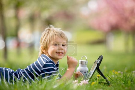 Téléchargez les photos : Belle enfant, garçon blond, jouant sur tablette dans le parc, bouteille d'eau à côté de lui - en image libre de droit