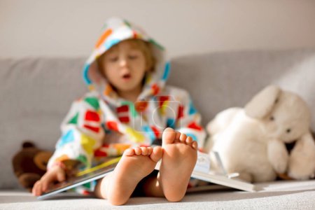 Photo for Happy toddler child, blond boy, sitting on the couch with stuffed toy after bath, reading a book, smiling ,colorful bathrobe - Royalty Free Image