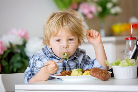 Téléchargez les photos : Petit enfant en bas âge, garçon blond, manger des légumes bouillis, brocoli, pommes de terre et carottes avec de la viande de poulet frite à la maison, tout en regardant un film sur tablette - en image libre de droit
