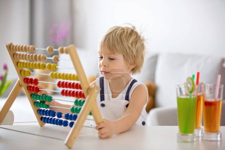 Photo for Sweet toddler child, blond boy, learning math at home with colorful abacus, drinking freshly made juice - Royalty Free Image