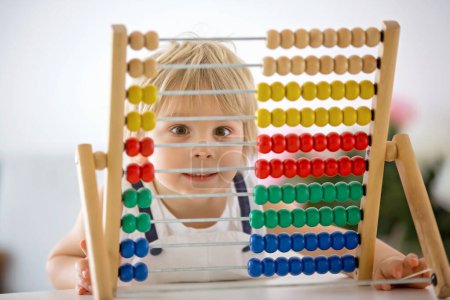 Sweet toddler child, blond boy, learning math at home with colorful abacus, drinking freshly made juice