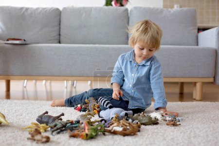 Foto de Lindo niño rubio, niño pequeño, jugando en casa con dinosaurios, sentado en el suelo - Imagen libre de derechos
