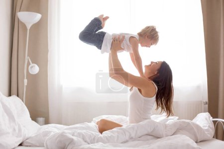 Foto de Hermoso niño rubio y su madre, jugando en casa por la mañana en la cama, sonriendo, riendo y divirtiéndose juntos - Imagen libre de derechos