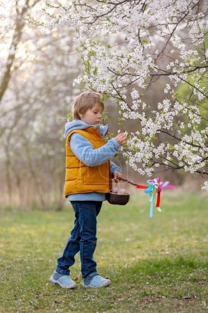 Photo for Beautiful blond child, boy, holding twig, braided whip made from pussy willow, traditional symbol of Czech Easter used for whipping girls and basket with eggs in park - Royalty Free Image