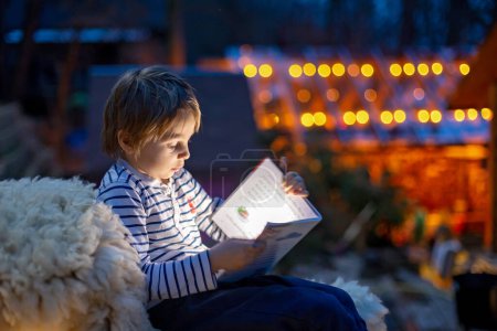Photo for Beautiful blond child, boy, reading a book at night with flashligh, sitting on the stairs in garden in cottage village house - Royalty Free Image
