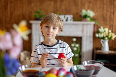 Foto de Beatiful niño rubio, niño, colorear y pintar huevos para Pascua en casa, preparándose para las vacaciones - Imagen libre de derechos