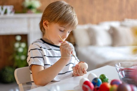 Téléchargez les photos : Enfant blond, garçon, colorant et peignant des œufs pour Pâques à la maison, se préparant pour les vacances - en image libre de droit