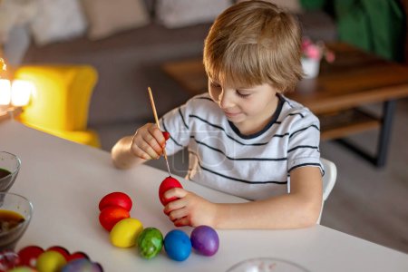 Téléchargez les photos : Enfant blond, garçon, colorant et peignant des œufs pour Pâques à la maison, se préparant pour les vacances - en image libre de droit