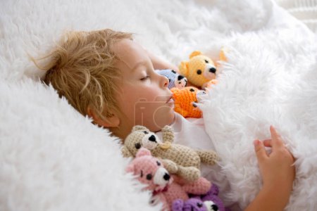Little toddler child, cute blond boy, sleeping with many teddy bears, handmade amigurumi toys at home in bed