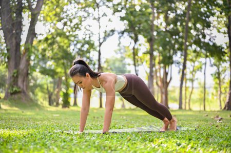 Foto de Hermosa y encantadora mujer asiática en ropa deportiva practicando pose de tablón completo, entrenando yoga en el parque natural verde. - Imagen libre de derechos