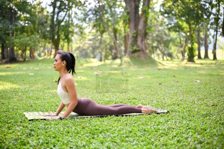 Attractive and healthy Asian woman in sportswear practicing yoga in the green park, doing Upward facing dog yoga pose on her mat.