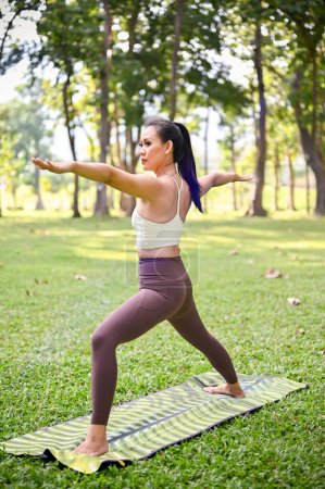 Asiatin in Sportbekleidung in Krieger-II-Yoga-Pose, praktiziert Yoga im schönen grünen Garten.